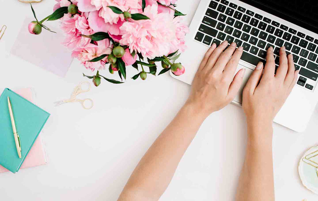 Close up of hands on a laptop keyboard next to a pink bouquet, a blue notebook, pen, paperclips, and scissors.